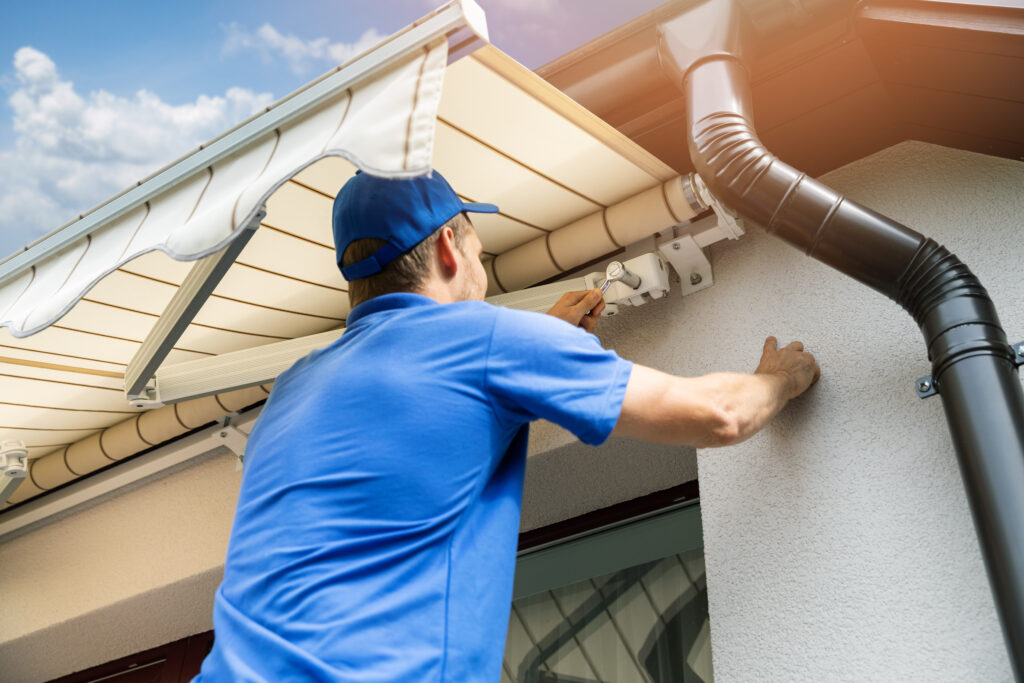 Ein Mann in blauer Kleidung befestigt eine gestreifte Markise an der Außenwand eines Hauses.