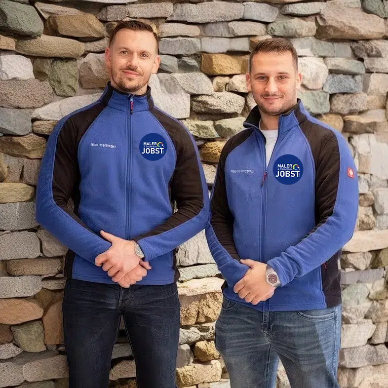 Die zwei Geschäftsführer in blau-schwarzen Jacken mit den Logos „MALER JOBST“ stehen selbstbewusst vor einer Steinmauer. Mit kurzen braunen Haaren und auf die Kamera gerichteten Augen strahlen sie Präzision und Engagement für Verbesserung aus. Maler & Trockenbau Jobst Lindau